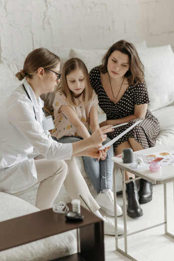 A doctor sits on a couch with a mother and daughter, showing them something on a clipboard.