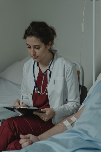 A doctor sits at the bedside of her patient, writing something on a clipboard