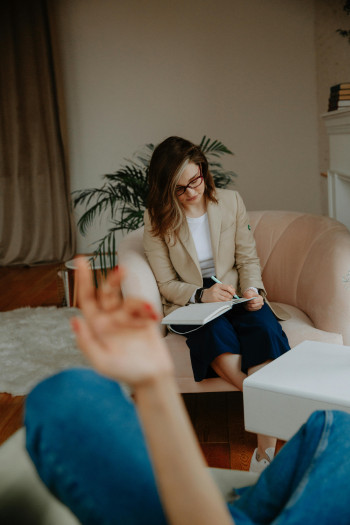 A woman wearing glasses takes notes as she talks to someone sitting across from her.