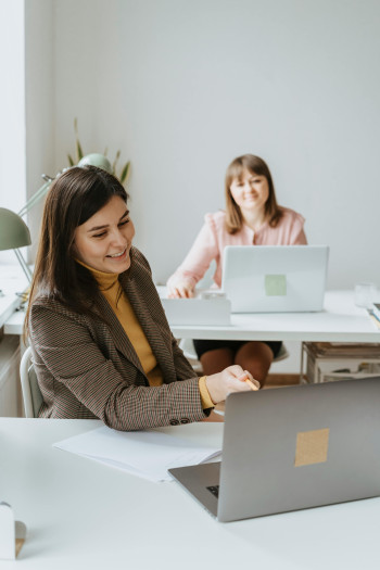 A woman working at her laptop points something out on the screen to her coworker at the desk behind her.