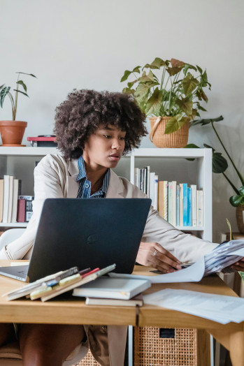 A black woman sits at her desk with her laptop. She is reaching over to a pile of paperwork.