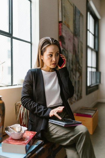 A woman sits on a bench with a tablet on her lap, talking on the phone. She has a serious expression on her face.