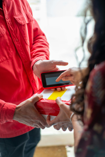 A person with long, curly hair accepts a parcel being delivered to them, and signs for it on a touchscreen device.