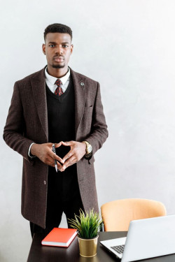 A black man stands looking at the camera. He is behind a desk with a laptop on it. He is wearing a black suit and tiw and a long brown coat. 