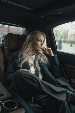 A businesswoman with long blonde hair sits in the back seat of a car looking out the window. She is wearing a loose dark suit with a white shirt. 
