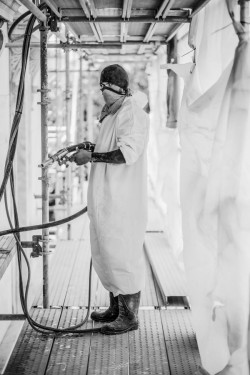 A person in protective gear stands in a draped worksite holding a sprayer. They are looking at the camera. 