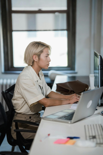 A person with short blonde hair sits at a desk, typing at a computer. There is an open laptop and post-it notes next to her on the desk.