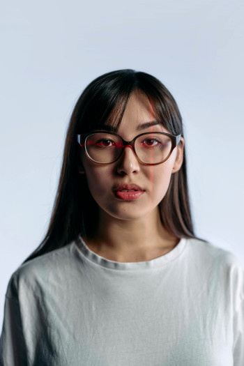 An Asian woman wearing glasses, shown from the shoulders up, looks directly at the camera. There is a red laser line running vertically down one side of her face, and another horizontally across her eyes, as though her face is being scanned.