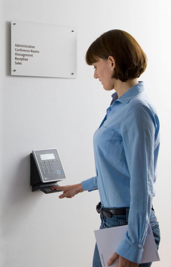 A woman stands and puts her finger into a fingerprint scanner to gain access to another space. She has a brown bobbed hairstyle with a fringe and wears a blue business shirt and blue jeans. She is carrying a notepad.