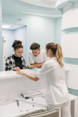 Two people stand at a reception desk in a medical facility, looking at a form on the counter. A person wearing white scrubs assists them.