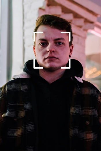 A white man with short, dark hair stands straight on to the camera. Four corners of a square surround his face as though being scanned by something.