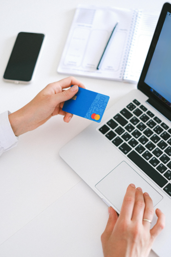 A person holds a bank card in one hand and uses a laptop with the other. A cell phone and notebook are in the background.
