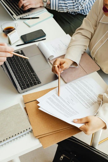 Three people do paperwork on a desk with documents and laptops all over it. Only their hands are visible, and they are gesturing at one set of papers in particular.
