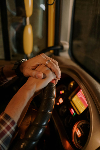 A person's hands rest on the top of a steering wheel of a bus or large vehicle. It is dark outside and lights on the dashboard are glowing.