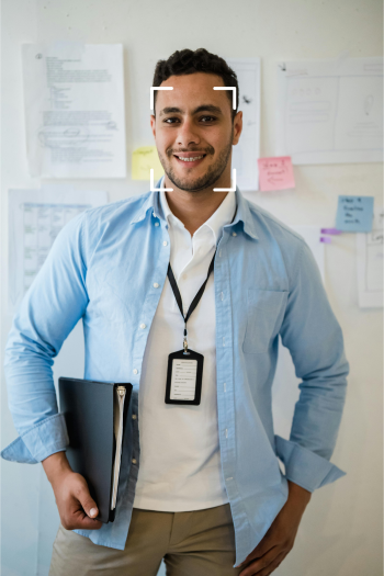 A man smiles at the camera while holding a folder of paperwork under one arm. He's wearing a blue button down shirt over a while polo, and has a lanyard around his neck.