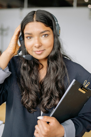 A woman wearing a headset and holding a clipboard smiles at the camera.