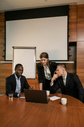 Three business people in dark suits at a table look at a laptop screen. Two are sitting and one stands.