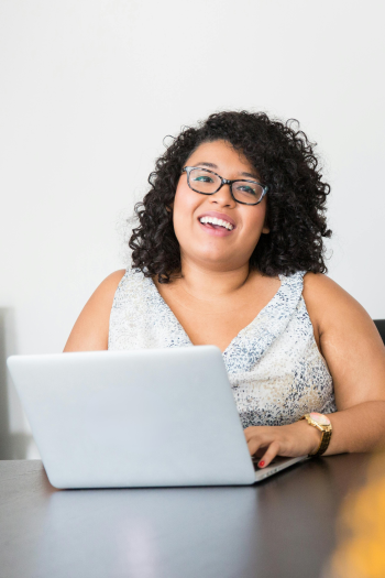 A curly dark-haired woman in a sleeveless top sits behind a laptop. She has glasses and is similing.