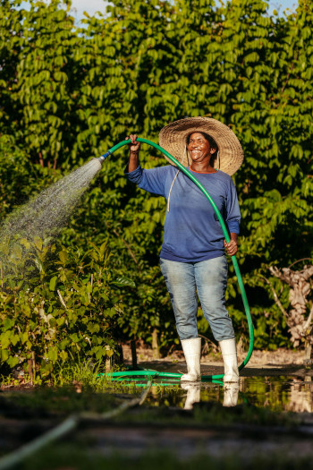 A black woman looks happy watering her garden with a hose. She is wearing tall white gumboots, jeans, and a blue top.