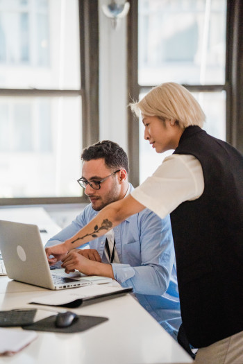 A person with blonde hair is standing behind a man sitting who is wearing glasses and a blue shirt. Both are looking at a laptop on a desk.
