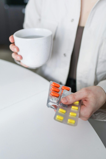 A person holds two blister packs of orange and yellow pills and a cup of water.