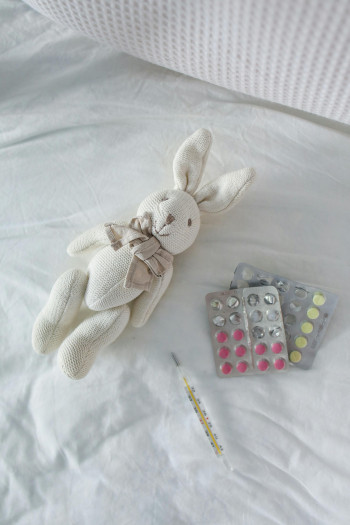 A white toy rabbit lies on a white sheet beside two pill packets and a thermometer.