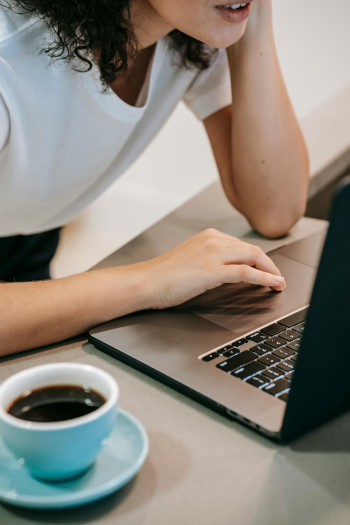 A woman in a white t-shirt hovers over a laptop looking focused. Her face is cropped so we can only see her mouth and lower face. A blue cup of coffee sits to the right of the laptop.