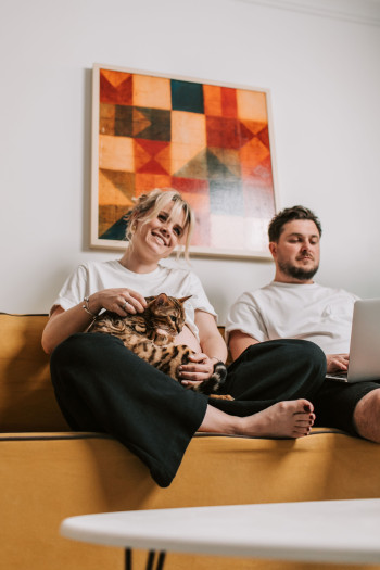 A white couple, a man and a woman, sit on the couch with a cat. They are wearing white t-shirts and black trousers. The man is on a laptop. 