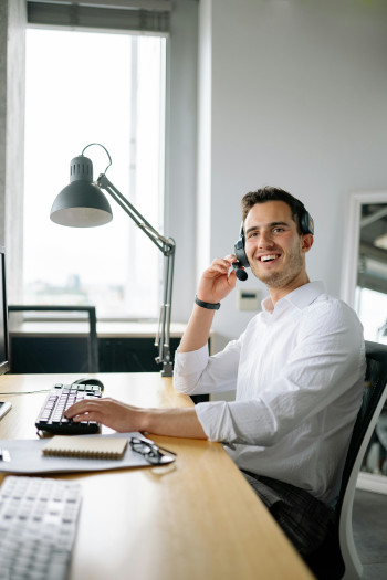 A man sits in an office at a computer. He has a phone headset on and looks into the distance, smiling. He is weraing a white collared shirt and has dark brown hair and face stubble.