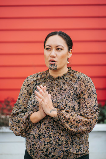 A woman in a light, brown top stands with her hands pressed together. She is talking and has a moko and dark hair pulled back. She is in front of a red wall.