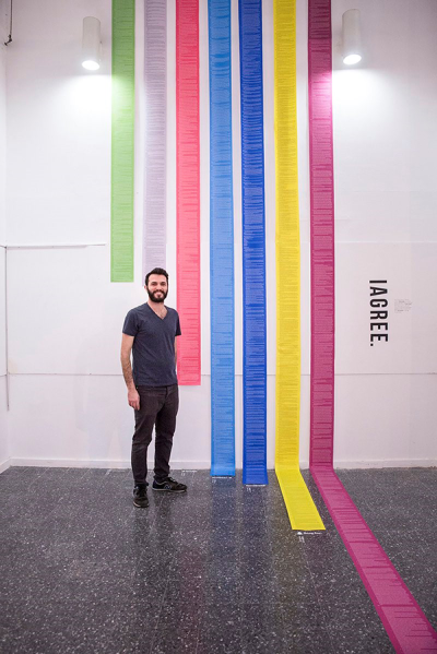 A dark-haired man with a beard stands in a gallery beside a white wall. He is wearing a grey t-shirt and black jeans. On the wall are coloured print outs of documents showing how long they are.
